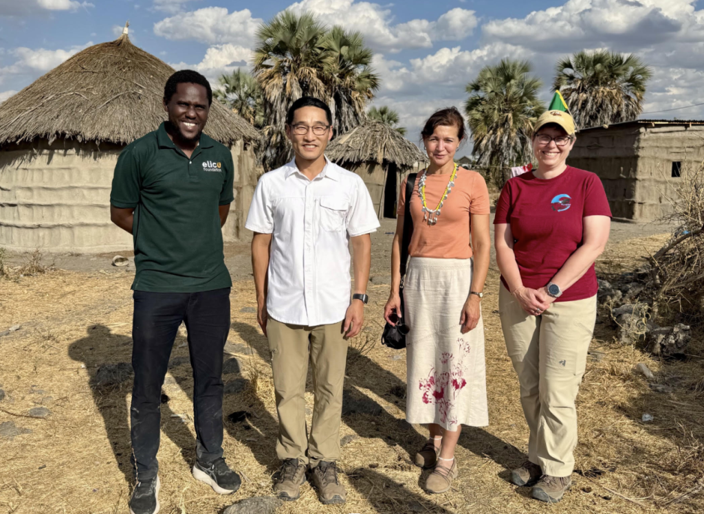 The Bold Challenges team stands in a rural village in front of a traditional round, thatched-roof homestead