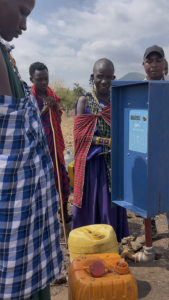 Villagers encircle a water faucet dispensing water into an orange utility jug
