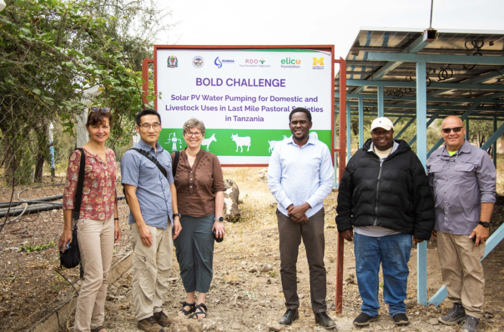 Team members pose in front of a framed sign that reads, "Bold Challenge: Solar PV Water Pumping for Domestic and Livestock Uses in Last Mile Pastoral Societies in Tanzania, with partner logos